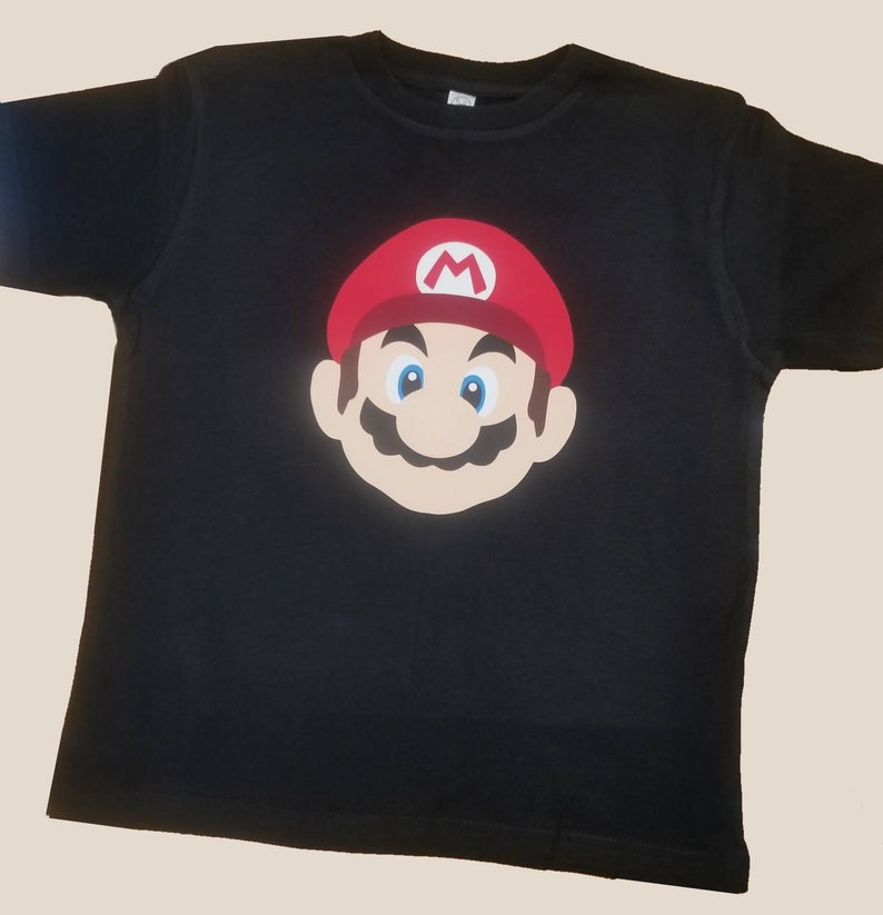 Op de afbeelding: Zwart T-shirt met een rode en witte Mario-personage afbeelding.