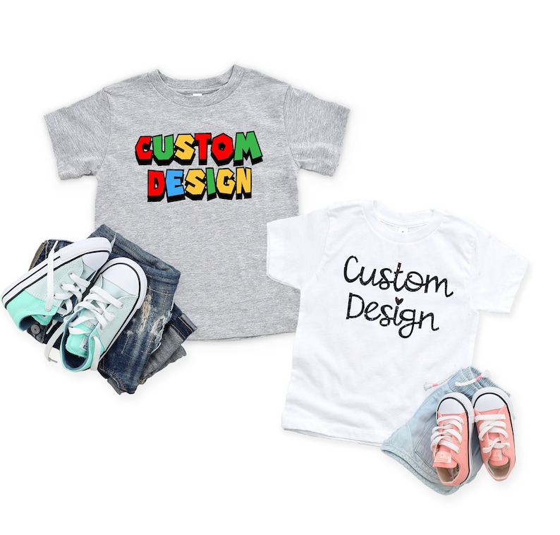 Op de afbeelding: Twee t-shirts in peutermaat met de tekst "Custom Design" erop. E&eacute;n shirt is grijs met de tekst in kleurrijke blokletters. Het andere shirt is wit met de tekst in zwarte cursieve letters.