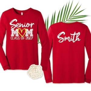 Op de afbeelding: Twee rode shirts met lange mouwen. De voorkant heeft witte tekst met Senior MOM, een geel hart en CLASS OF 2027. De achterkant toont de naam Smith in witte schrijfletters. Een witte bloem en een groen palmblad liggen tussen de shirts in.