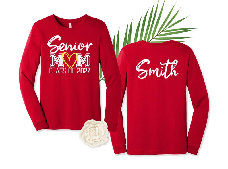 Op de afbeelding: Twee rode shirts met lange mouwen. De voorkant heeft witte tekst met Senior MOM, een geel hart en CLASS OF 2027. De achterkant toont de naam Smith in witte schrijfletters. Een witte bloem en een groen palmblad liggen tussen de shirts in.