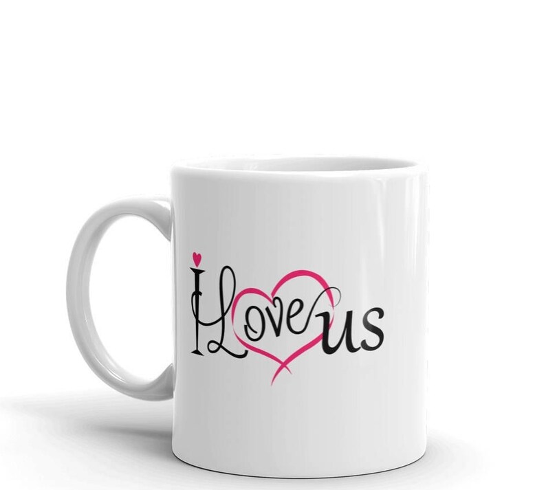 May include: White ceramic mug with the words "I Love Us" in black script and a pink heart design. The mug has a curved handle and is set against a white background.
