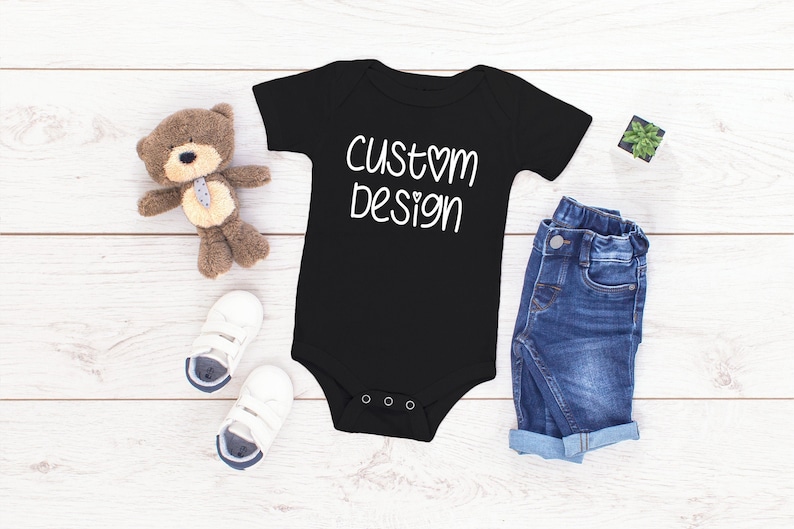 May include: Black baby onesie with white text that says "custom design" with a heart symbol. The onesie is laid out on a white wooden surface with a teddy bear, a pair of baby shoes, and a small potted plant.