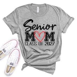 Op de afbeelding: Een grijs gem&ecirc;leerd T-shirt met korte mouwen en de zwarte tekst Senior MOM, waarbij een handgetekend rood hart de letter O vervangt, en de zwarte tekst CLASS OF 2027 eronder. Het shirt heeft een ronde hals en een geknoopt detail aan de zoom.