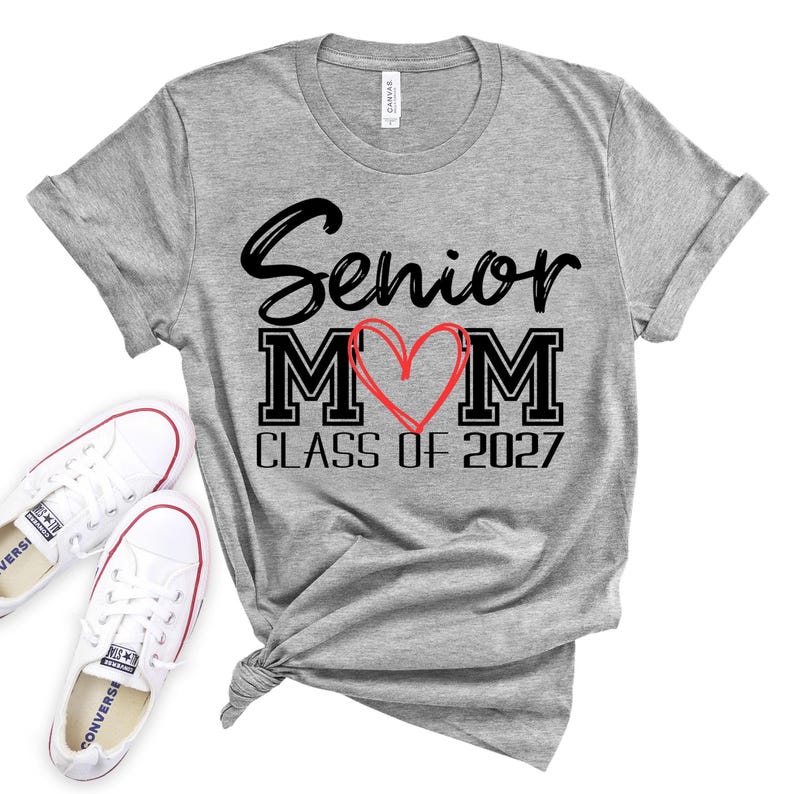 Op de afbeelding: Een grijs gem&ecirc;leerd T-shirt met korte mouwen en de zwarte tekst Senior MOM, waarbij een handgetekend rood hart de letter O vervangt, en de zwarte tekst CLASS OF 2027 eronder. Het shirt heeft een ronde hals en een geknoopt detail aan de zoom.