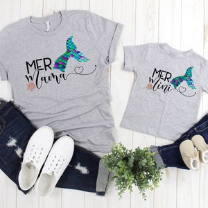 May include: Two gray t-shirts with a mermaid tail graphic and the text "Mer Mama" and "Mer Mini". The shirts are laid out on a white wooden surface with jeans, shoes, and a potted plant.