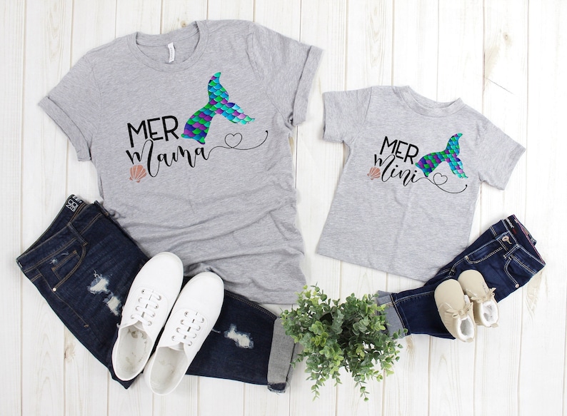 May include: Two gray t-shirts with a mermaid tail graphic and the text "Mer Mama" and "Mer Mini". The shirts are laid out on a white wooden surface with jeans, shoes, and a potted plant.