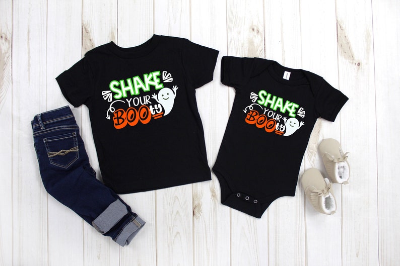 May include: Two black shirts with the text "Shake Your Boo-ty" and a ghost graphic. The shirts are on a white wooden background. One shirt is a long-sleeved t-shirt and the other is a short-sleeved onesie.