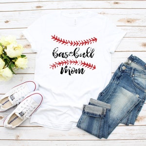 May include: White t-shirt with the text "baseball mom" in black lettering and red baseball stitching. The shirt is laid out on a white wood background with a pair of white sneakers and blue jeans.