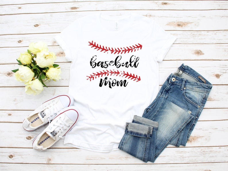 May include: White t-shirt with the text "baseball mom" in black lettering and red baseball stitching. The shirt is laid out on a white wood background with a pair of white sneakers and blue jeans.