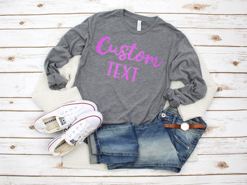 May include: A gray long-sleeved t-shirt with the text "Custom Text" in pink glitter lettering. The shirt is laid out on a white wooden surface with a pair of white sneakers, blue jeans, and a white cardigan.