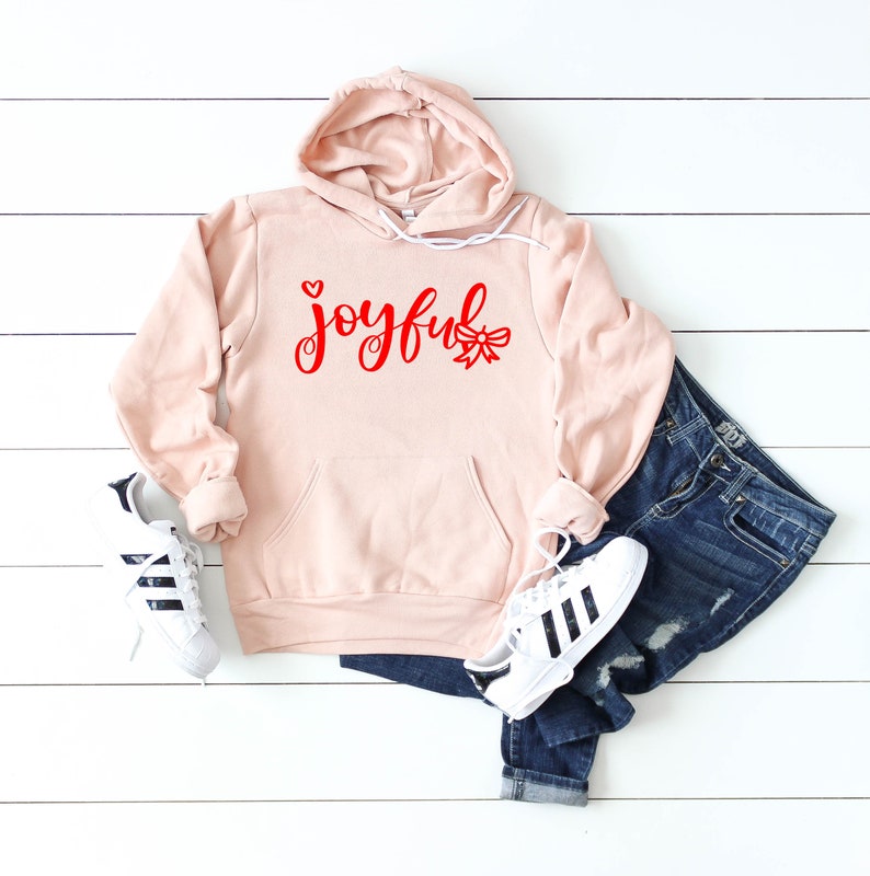 May include: A pink hoodie with a red "joyful" graphic with a heart and a bow. The hoodie is laid out on a white wooden surface with a pair of black and white sneakers and blue jeans.