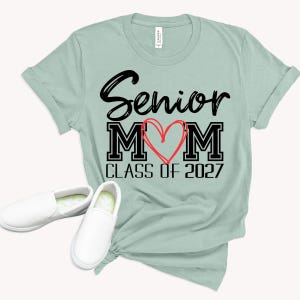 Op de afbeelding: Een licht saliegroen t-shirt met korte mouwen met de zwarte tekst Senior MOM, een rood handgetekend hart in het midden en de zwarte tekst CLASS OF 2027 eronder. Twee witte instapschoenen zijn bij de onderkant van het t-shirt geplaatst.