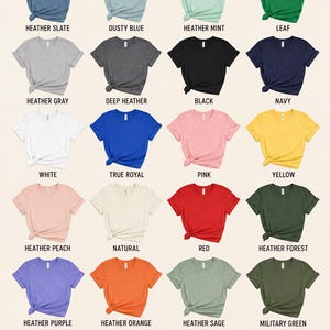 Op de afbeelding: Een kleurenkaart met twintig t-shirts met korte mouwen in verschillende tinten, waaronder heather slate, dusty blue, heather mint, leaf, heather gray, deep heather, black, navy, white, true royal, pink, yellow, heather peach, natural, red, heather forest, heather purple, heather orange, heather sage en military green, met de tekst T-SHIRT COLOR OPTIONS bovenaan.