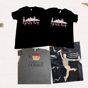 Puede incluir: Tres camisetas con diferentes dise&ntilde;os. Las dos primeras son negras con un horizonte de ciudad blanco y el texto "Girls Trip" en rosa. La tercera es gris con una taza de caf&eacute; de dibujos animados con una cara y el texto "Espresso Patronum".
