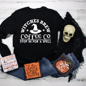 May include: Black long-sleeved t-shirt with white text that reads "Witches Brew Coffee Co. Stop in for a spell". The text is in a circle with a witch's hat and stars. The shirt is laying on a white wooden surface with a pair of blue jeans, a skull, and three Halloween-themed signs.