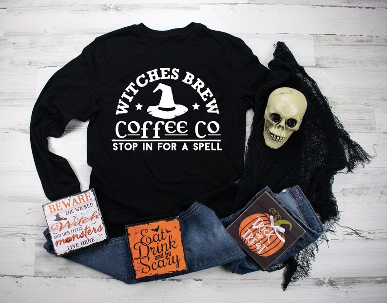May include: Black long-sleeved t-shirt with white text that reads "Witches Brew Coffee Co. Stop in for a spell". The text is in a circle with a witch's hat and stars. The shirt is laying on a white wooden surface with a pair of blue jeans, a skull, and three Halloween-themed signs.