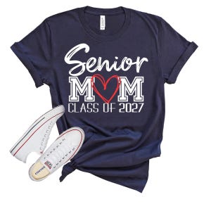 Op de afbeelding: Een marineblauw t-shirt met korte mouwen en witte tekst Senior MOM, waarbij een rood hart de letter O vervangt, en de tekst CLASS OF 2027 eronder. Twee witte canvas sneakers met zwarte en rode randen staan aan de linkerkant.