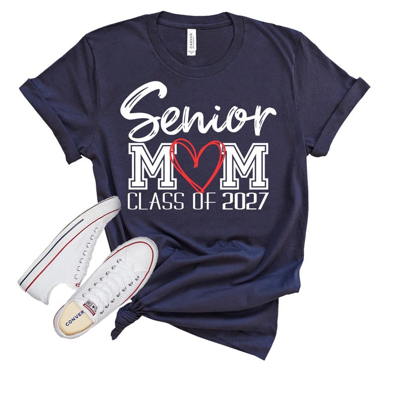 Op de afbeelding: Een marineblauw t-shirt met korte mouwen en witte tekst Senior MOM, waarbij een rood hart de letter O vervangt, en de tekst CLASS OF 2027 eronder. Twee witte canvas sneakers met zwarte en rode randen staan aan de linkerkant.