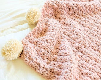 May include: A chunky crochet blanket in a soft dusty pink colour featuring a textured stitch pattern. The edges of the blanket are decorated with several large, round, cream-coloured yarn pom-poms attached at regular intervals along the border.