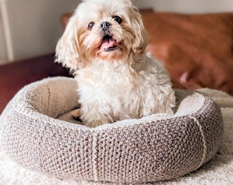May include: A small, white dog with brown eyes sits in a light brown crocheted dog bed. The dog is smiling and has its tongue sticking out.
