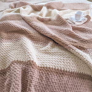 May include: A soft, striped crocheted throw blanket in shades of beige, brown, and pink. The blanket is folded and draped over a bed.