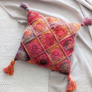 May include: A crocheted pillow with a square pattern in shades of pink, orange, and brown. The pillow has tassels on each corner.