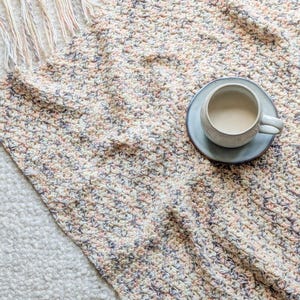 May include: A handmade crochet blanket in a speckled pattern of cream, pink, and gray. The blanket has a fringe edge and a cup of coffee on a blue saucer. The blanket is on a white surface.