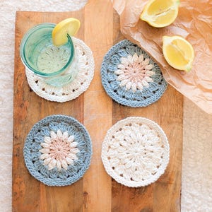 Może przedstawiać: Cztery ręcznie robione podkładki z szydełka w kolorze białym i niebieskim z kwiatowym wzorem. Jedna podkładka zawiera szklankę wody z plasterkiem cytryny. Podkładki znajdują się na drewnianej desce do krojenia z dwiema połówkami cytryny.