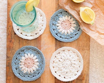 Pode incluir: Quatro bases de crochet feitas à mão em branco e azul com um design floral. Uma base contém um copo de água com uma fatia de limão. As bases estão numa tábua de cortar de madeira com duas metades de limão.
