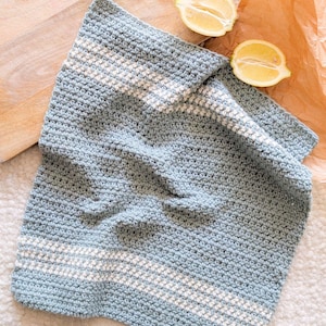 May include: A light blue crocheted dishcloth with white stripes. The dishcloth is folded and lying on a white surface.