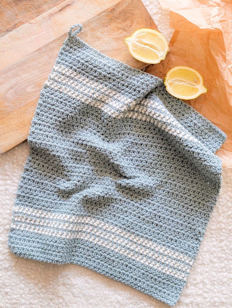 Puede incluir: Un pa&ntilde;o de cocina de crochet azul claro con rayas blancas. El pa&ntilde;o de cocina est&aacute; doblado y sobre una superficie blanca.