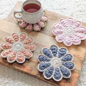 May include: A collection of handmade crochet flower coasters in various colors, including pink, peach, and blue. One coaster supports a white mug filled with tea. The coasters are arranged on a wooden board, showcasing their intricate floral design.