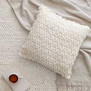 Puede incluir: Un cojín de tiro de crochet de color crema con una superficie texturizada se encuentra sobre una alfombra texturizada blanca y beige.