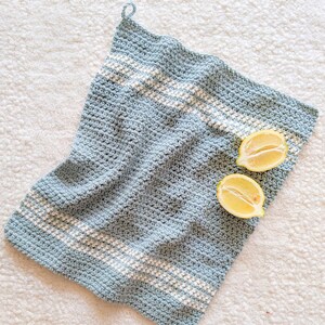 Puede incluir: Un pa&ntilde;o de cocina de crochet azul claro con rayas blancas. La toalla est&aacute; colgada en una superficie blanca con dos mitades de lim&oacute;n encima.