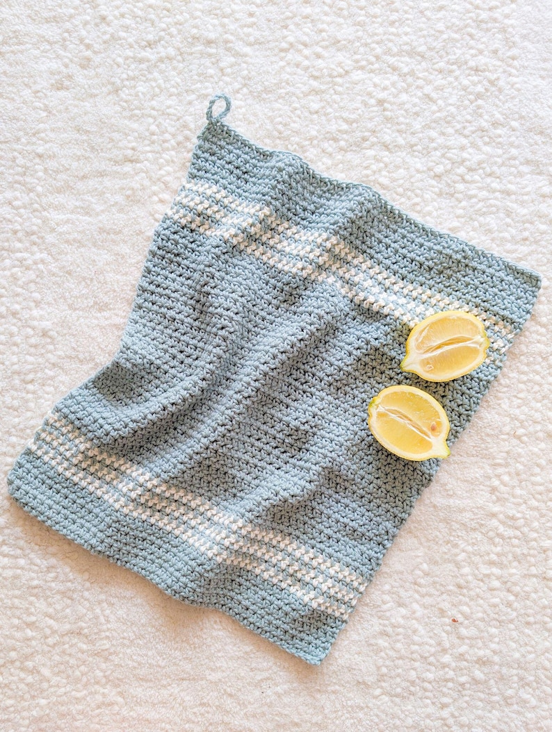 Puede incluir: Un pa&ntilde;o de cocina de crochet azul claro con rayas blancas. La toalla est&aacute; colgada en una superficie blanca con dos mitades de lim&oacute;n encima.