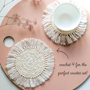 May include: Two white crocheted coasters with fringe, one with a cup on top. The text "crochet 4 for the perfect coaster set!" is written on the pink surface.