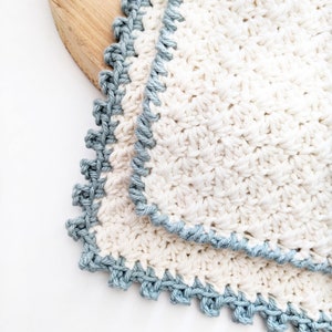 May include: Two crocheted dishcloths with a blue trim. The dishcloths are made of a white cotton yarn and have a textured pattern.