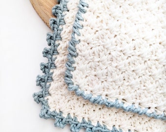 May include: Two crocheted dishcloths with a blue trim. The dishcloths are made of a white cotton yarn and have a textured pattern.