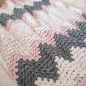 Op de afbeelding: Een gehaakt deken met een chevron-patroon in tinten roze, grijs en wit.