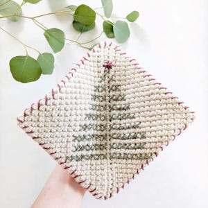 May include: A white knitted dishcloth with a green Christmas tree design and a burgundy border.