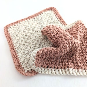 May include: Two knitted dishcloths, one in a light beige color and the other in a dusty rose color. The dishcloths are made with a simple stitch pattern and have a crocheted border.
