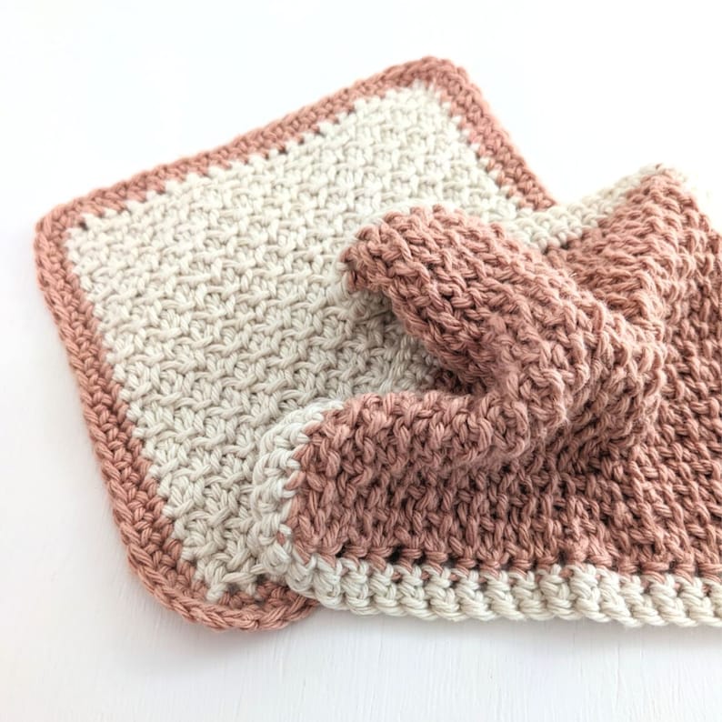 May include: Two knitted dishcloths, one in a light beige color and the other in a dusty rose color. The dishcloths are made with a simple stitch pattern and have a crocheted border.