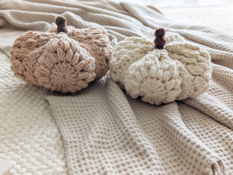 Op de afbeelding: Twee handgehaakte pompoenen, &eacute;&eacute;n in lichtbeige en de andere in off-white. Elke pompoen heeft een gestructureerd patroon van pofsteken en een kleine, donkerbruine steel aan de bovenkant, wat zorgt voor een ronde, zachte en decoratieve uitstraling.
