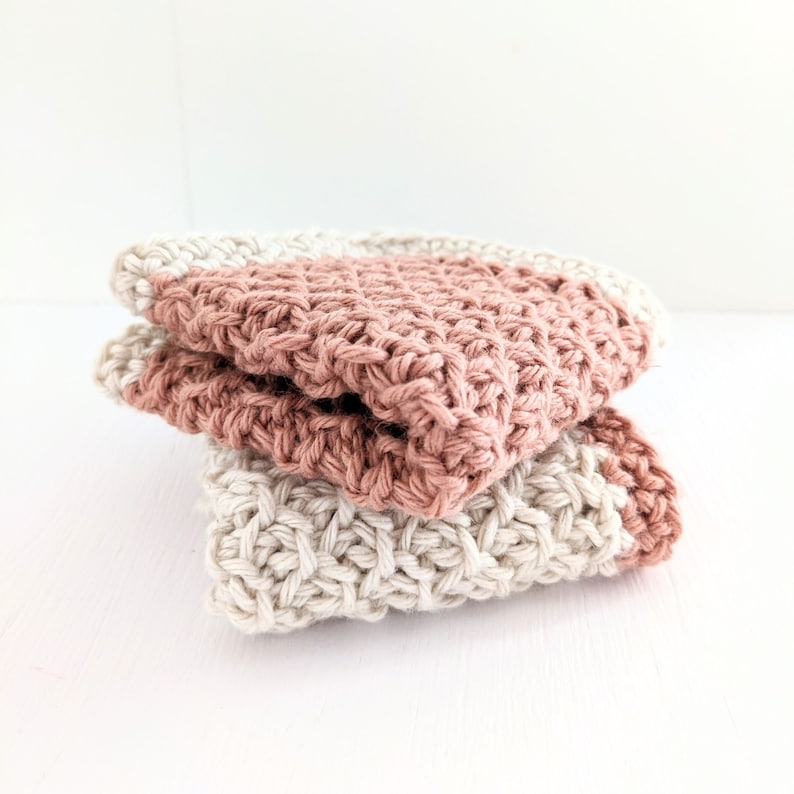 May include: Two crocheted dishcloths, one in a light beige color and the other in a dusty rose color. The dishcloths are folded and stacked on top of each other.