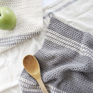 Puede incluir: Dos pa&ntilde;os de cocina de ganchillo, uno blanco y otro gris con rayas blancas. El pa&ntilde;o de cocina gris est&aacute; doblado y una cuchara de madera est&aacute; encima.