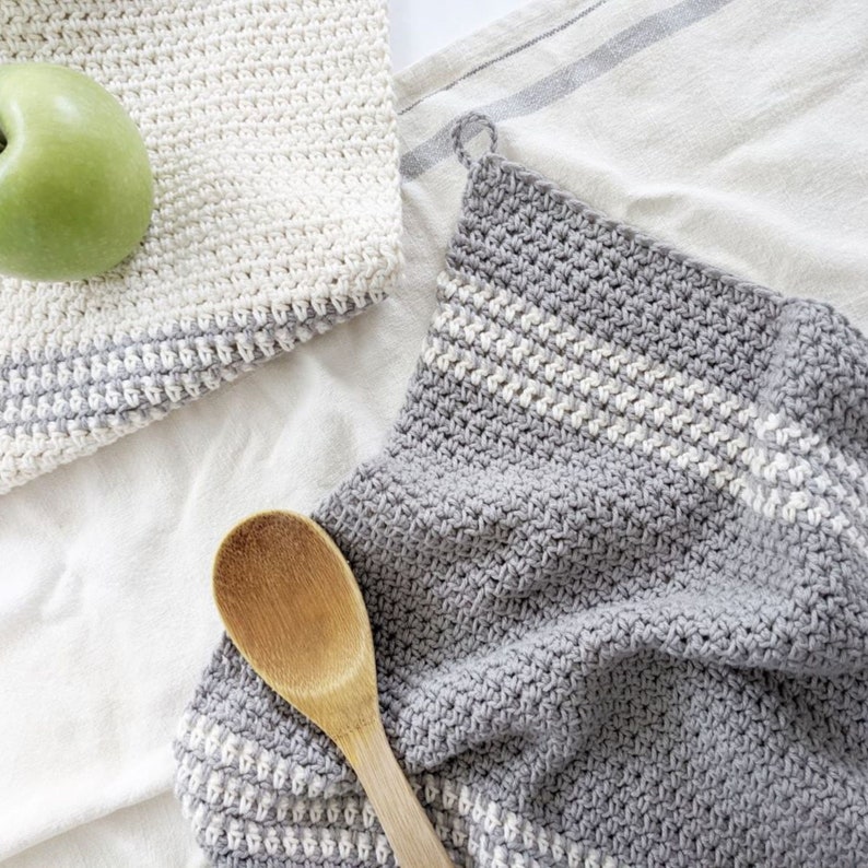 Puede incluir: Dos pa&ntilde;os de cocina de ganchillo, uno blanco y otro gris con rayas blancas. El pa&ntilde;o de cocina gris est&aacute; doblado y una cuchara de madera est&aacute; encima.
