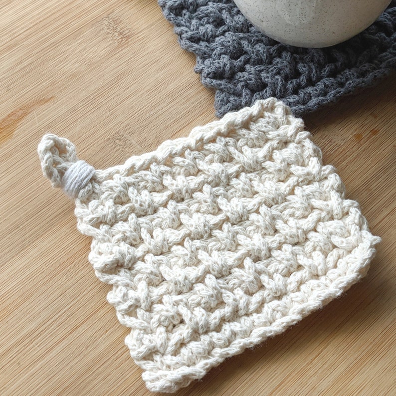 May include: A white crocheted square coaster with a knotted loop handle. The coaster is made with a textured stitch pattern.
