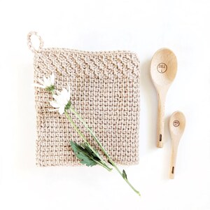 May include: A beige crocheted pot holder with a white flower and green leaves on top of it. Two wooden spoons with the words "1 TBS" and "1/2 tsp" are on a white surface.