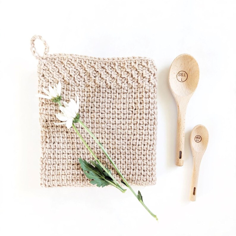 May include: A beige crocheted pot holder with a white flower and green leaves on top of it. Two wooden spoons with the words "1 TBS" and "1/2 tsp" are on a white surface.