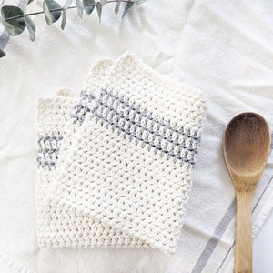 Puede incluir: Dos pa&ntilde;os de cocina de crochet blanco con rayas grises. Los pa&ntilde;os est&aacute;n doblados y colocados sobre una superficie blanca con una cuchara de madera.